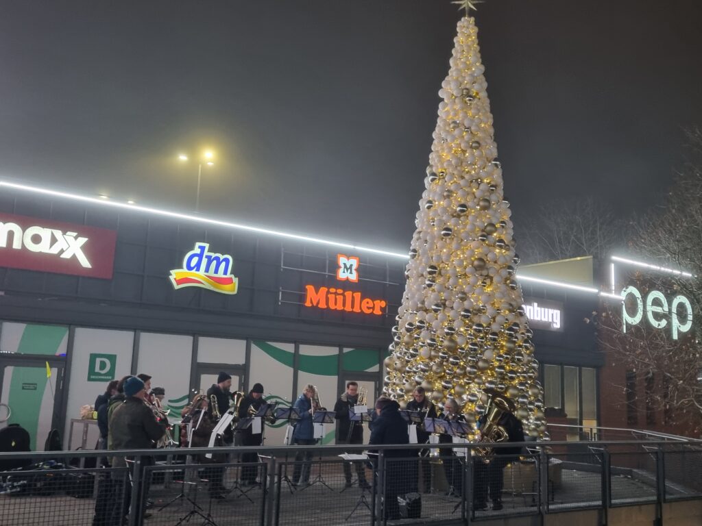 Eine Gruppe von Musikern spielt Blechblasinstrumente vor einem Einkaufszentrum, das nachts mit einem großen beleuchteten Weihnachtsbaum geschmückt ist. Die Schaufenster hinter ihnen sind beleuchtet und weisen verschiedene Ladenschilder auf.
