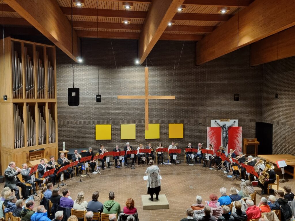 Ein Dirigent leitet eine Blaskapelle, die in einem Halbkreis in einer Kirche mit einem großen Holzkreuz an einer Backsteinmauer aufgestellt ist. Das Publikum schaut sitzend zu. Die Szene ist mit Holzbalken an der Decke warm beleuchtet.