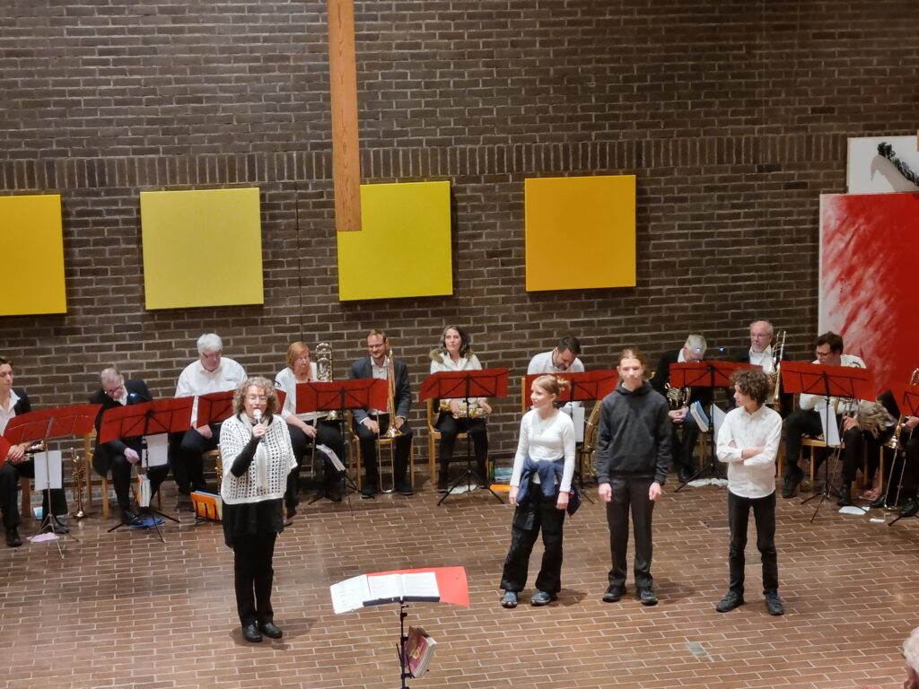 Eine Frau spricht auf der Bühne neben drei Personen in ein Mikrofon, dahinter sitzt eine Gruppe von Musikern mit Instrumenten und roten Notenständern vor einer Backsteinwand mit gelben Tafeln.