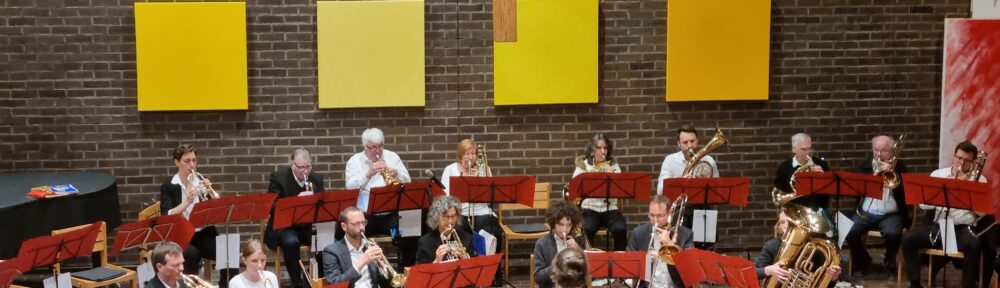 Ein Dirigent leitet ein Blechbläserensemble, das in Reihen sitzt und in einem gemauerten Raum mit einem großen Holzkreuz und gelben Tafeln an der Wand Instrumente spielt. Im Vordergrund sind Zuhörer zu sehen.