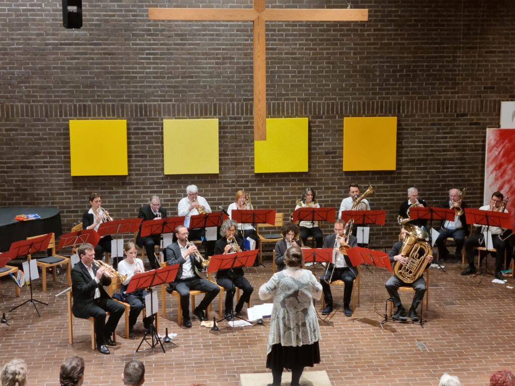 Ein Dirigent leitet ein Blechbläserensemble, das in Reihen sitzt und in einem gemauerten Raum mit einem großen Holzkreuz und gelben Tafeln an der Wand Instrumente spielt. Im Vordergrund sind Zuhörer zu sehen.