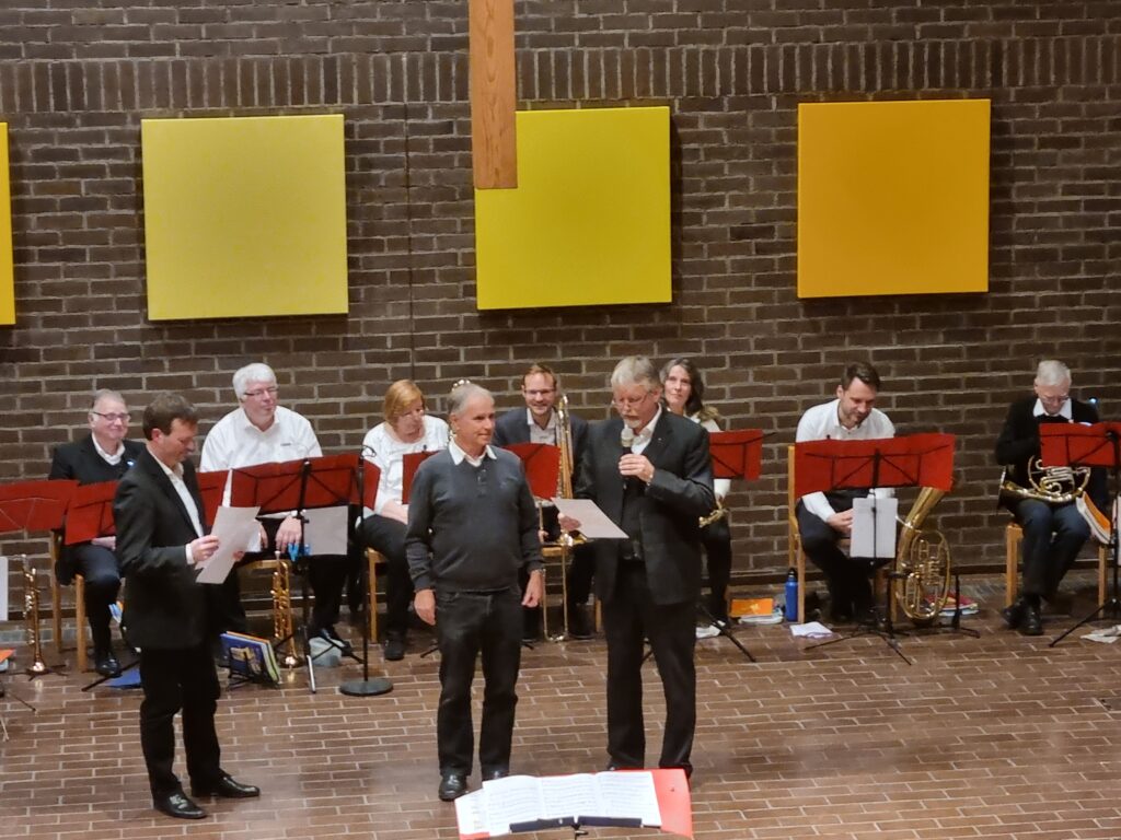 Eine Gruppe von Musikern mit Blech- und Holzblasinstrumenten sitzt auf Stühlen vor einer Backsteinwand mit gelben Platten. Drei Männer stehen davor, einer spricht in ein Mikrofon, während die anderen zuhören.