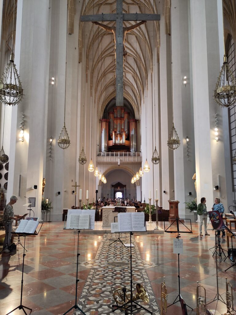 Ein Blick vom Altar einer prächtigen Kathedrale mit hohen Gewölbedecken, einem großen hängenden Kreuz, einer Orgel im hinteren Bereich, Kronleuchtern, Notenständern und im Vordergrund aufgestellten Blechblasinstrumenten.