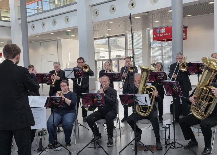 Eine Blaskapelle mit Musikern, die in einem modernen Innenraum sitzen, spielt Instrumente, darunter Tuben und Posaunen, während ein Dirigent vor ihnen steht. Die Notenständer haben bunte Hüllen.