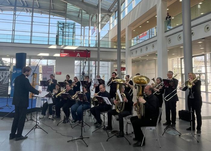 In einem hellen, modernen Gebäude mit Glaswänden und hohen Decken spielt eine große Blaskapelle, die von einem Dirigenten geleitet wird. Die Bandmitglieder sitzen und spielen verschiedene Blechblasinstrumente.