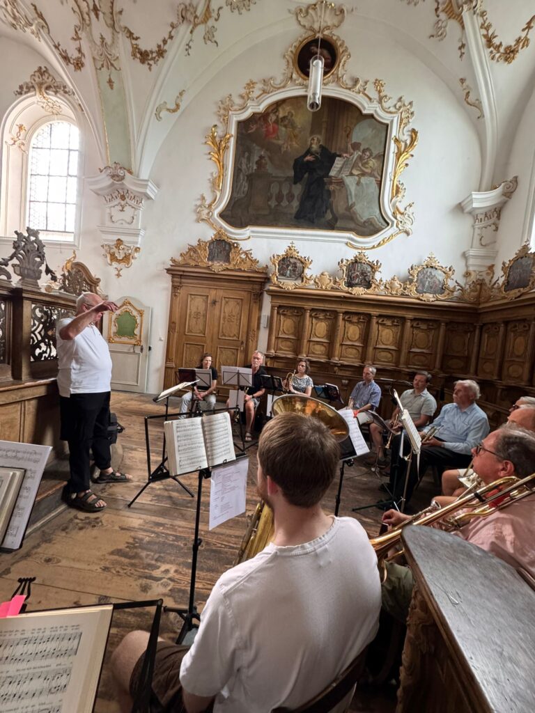 Eine Gruppe von Musikern probt in einem prunkvollen, historischen Raum mit Holzvertäfelung und einem großen Gemälde an der Wand. Vor ihnen steht ein Dirigent, der das Ensemble leitet. Noten und Notenständer sind zu sehen.