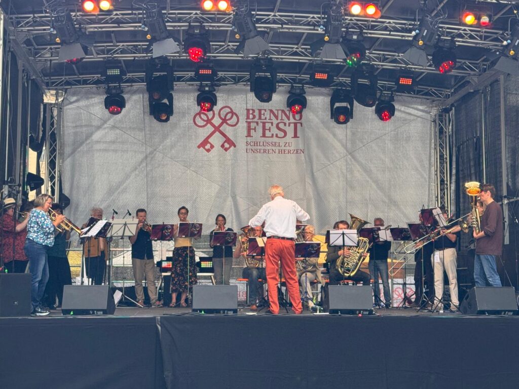 Ein Dirigent leitet eine Blaskapelle, die auf einer Freiluftbühne beim Benn Fest auftritt. Die Musiker spielen verschiedene Blechblasinstrumente, und hinter ihnen hängt ein Banner mit dem Namen der Veranstaltung. Bühnenlicht beleuchtet die Szene.