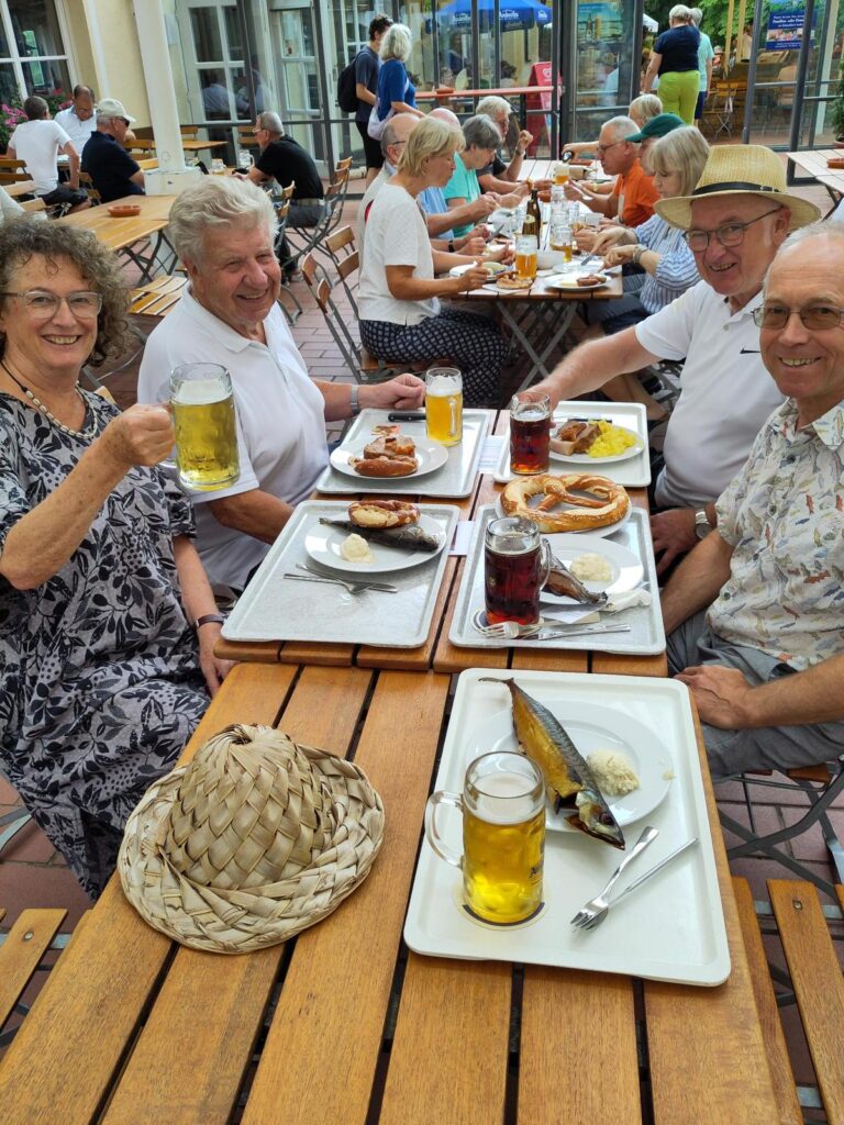 Vier ältere Menschen sitzen an einem hölzernen Tisch im Freien, lächeln und genießen Getränke sowie traditionelle deutsche Speisen. Zwei Männer und eine Frau halten Bierkrüge in der Hand; auf Tabletts stehen Teller mit Fisch, Wurst, Brezeln und Käse. In der Nähe sitzen weitere Gäste.