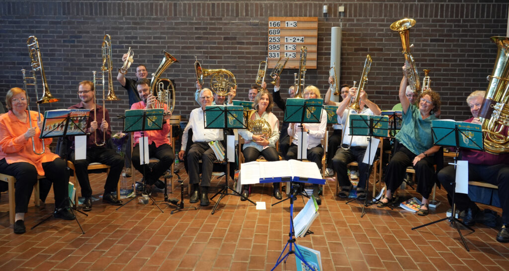 Eine Blaskapelle mit zwölf Musikern, die in zwei Reihen in einem Raum sitzen, halten ihre Instrumente hoch und lächeln in die Kamera. Vor ihnen stehen Notenständer, und im Hintergrund ist eine Backsteinwand mit Liednummern zu sehen.