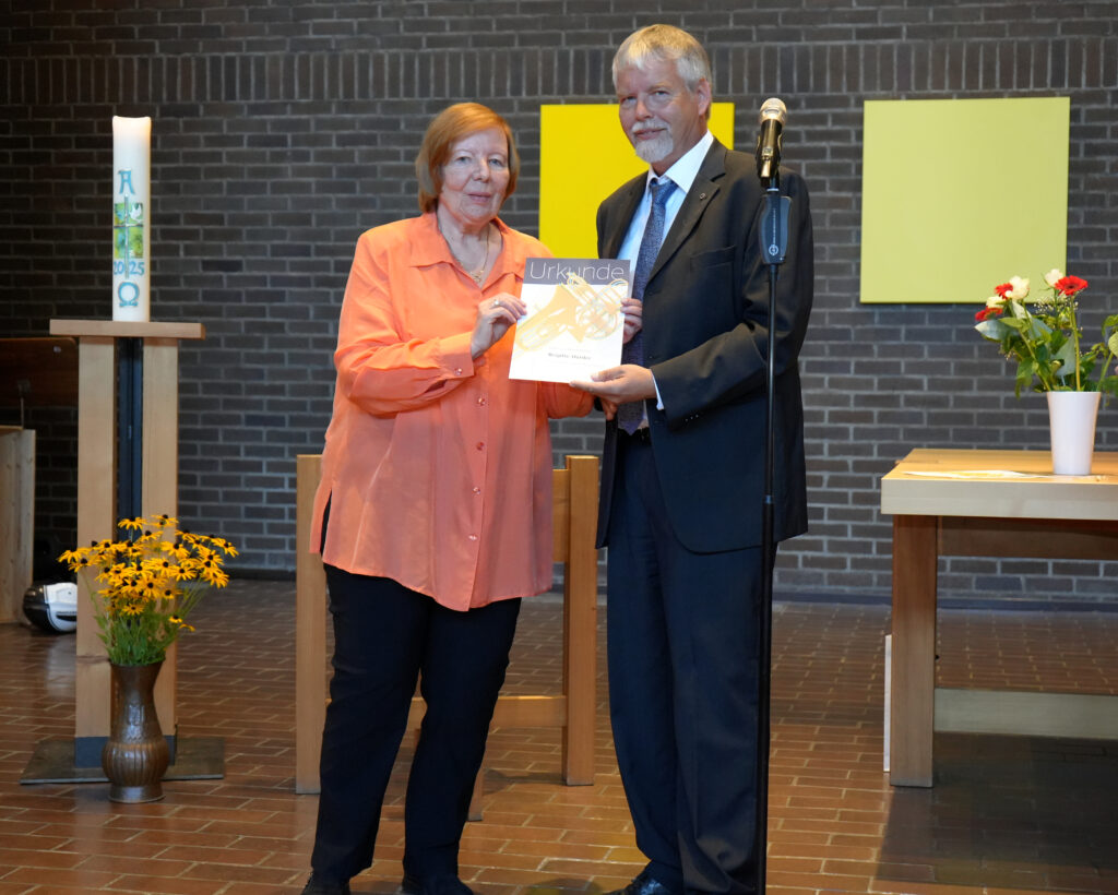Eine Frau in einem orangefarbenen Hemd und ein Mann in einem Anzug stehen nebeneinander in einer Kirche und halten eine Urkunde in der Hand. Sie lächeln beide. Ein Mikrofon, Blumen und Kerzen sind auf Holztischen hinter ihnen zu sehen.