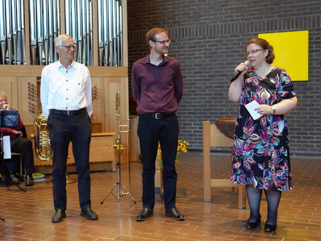 Drei Personen stehen in einem Raum vor Musikinstrumenten; eine Frau auf der rechten Seite spricht in ein Mikrofon, während zwei Männer links neben ihr stehen und zuhören. Im Hintergrund sind eine Backsteinmauer und eine Pfeifenorgel zu sehen.