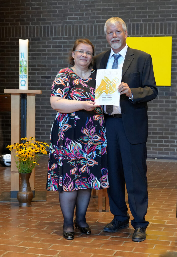 Eine Frau in einem farbenfrohen Kleid und ein Mann im Anzug stehen in einem Haus, lächeln und halten eine Urkunde in der Hand. Hinter ihnen stehen eine brennende Kerze, gelbe Blumen und eine gelbe rechteckige Dekoration an einer Backsteinmauer.