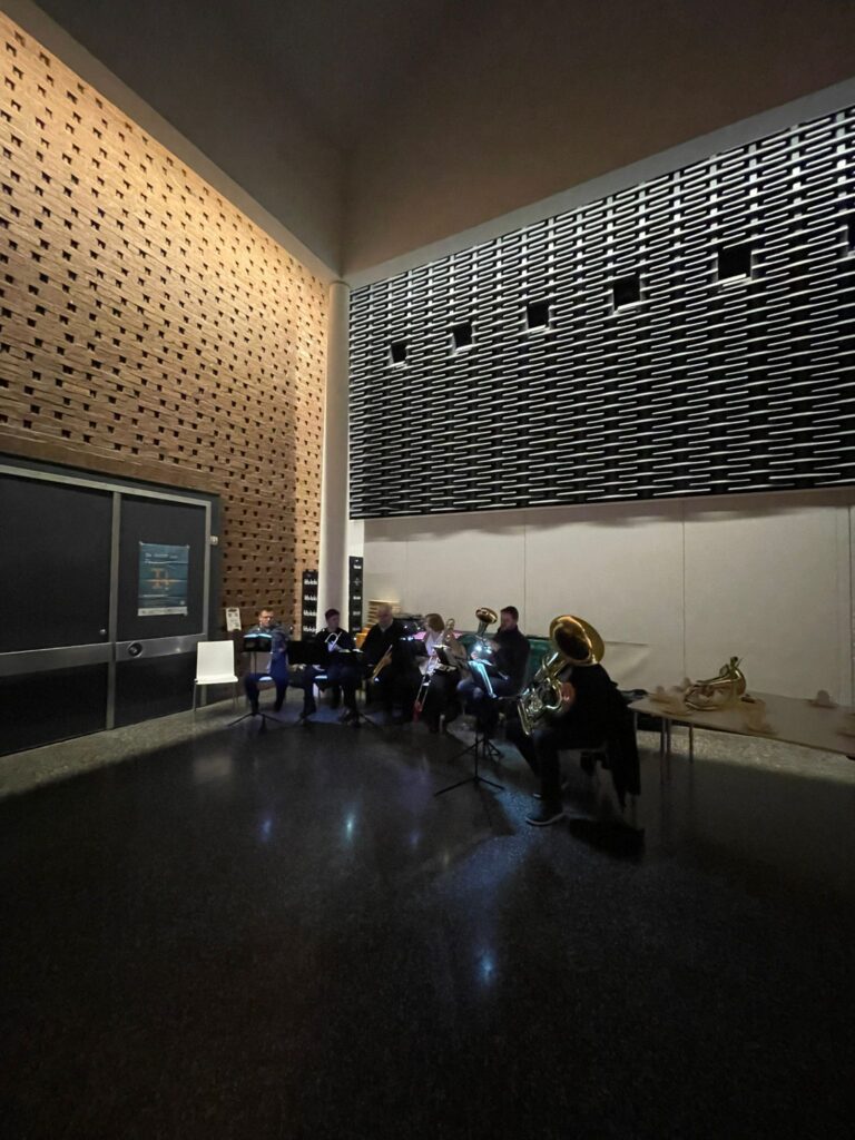 Eine kleine Blaskapelle spielt in einem schwach beleuchteten, modernen Innenraum mit hohen Backstein- und Metallwänden; Instrumente und Notenständer sind bei gedämpftem Licht sichtbar.