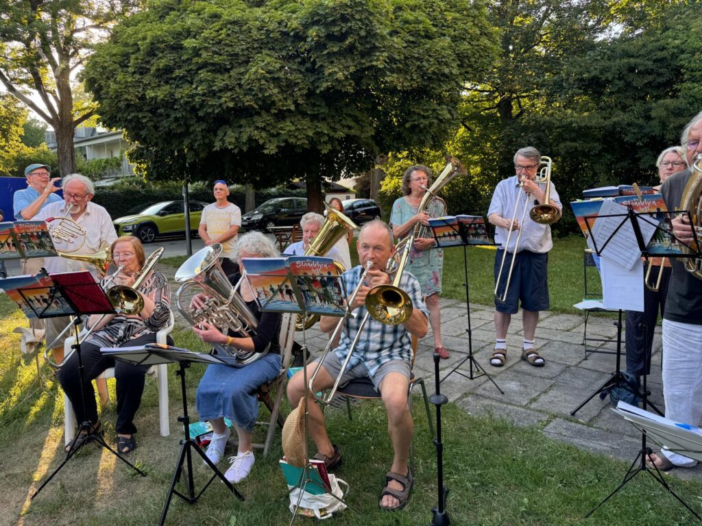Eine Gruppe von Menschen unterschiedlichen Alters spielt an einem sonnigen Tag im Freien Blechblasinstrumente; sie sitzen und stehen auf Gras und Pflaster, im Hintergrund sind Notenständer und Bäume zu sehen.