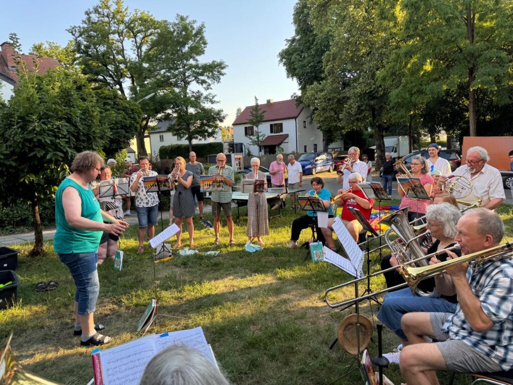 Eine Gruppe von Menschen, die an einem sonnigen Tag im Freien Blechblasinstrumente spielen, angeführt von einer Frau. Sie stehen und sitzen auf einer von Bäumen und Häusern umgebenen Rasenfläche, vor sich Notenständer.
