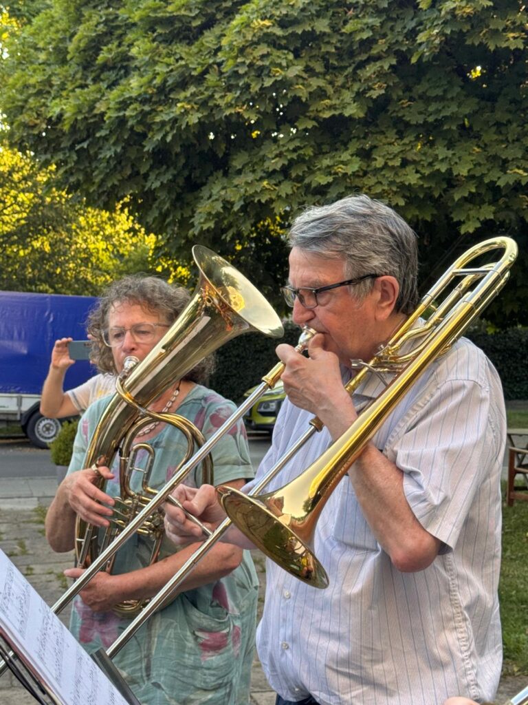 Zwei ältere Menschen spielen im Freien Blechblasinstrumente und lesen dabei Noten. Der Mann vorne spielt Posaune, die Frau dahinter Euphonium. Im Hintergrund sind Bäume, ein blauer Transporter und Sonnenlicht zu sehen.