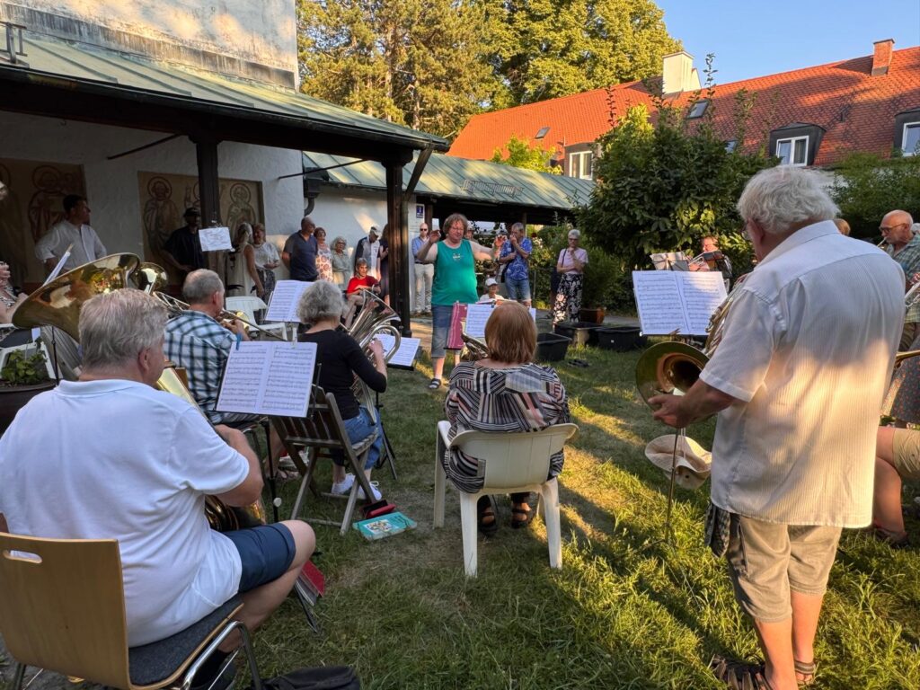 An einem sonnigen Tag spielt eine Blaskapelle im Freien vor einem sitzenden Publikum. Die Menschen haben sich in einem Garten versammelt; man sieht Notenständer, Instrumente und Notenblätter. Im Hintergrund sind Bäume und Gebäude zu sehen.