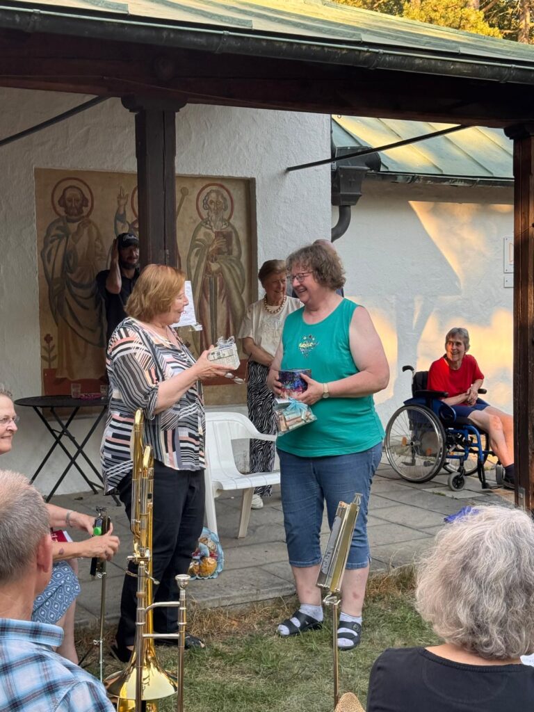Zwei Frauen stehen lächelnd vor einem Gebäude und tauschen Geschenke aus. Um sie herum haben sich mehrere Menschen versammelt, darunter eine Person im Rollstuhl, und im Vordergrund sind Blechblasinstrumente zu sehen. An der Wand hinter ihnen hängen religiöse Kunstwerke.