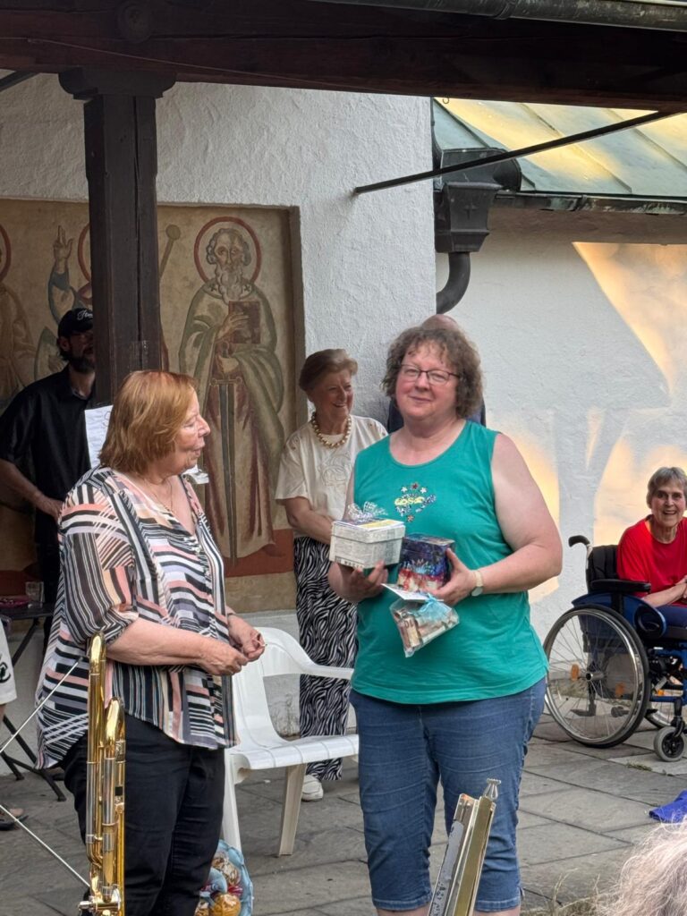Zwei Frauen stehen lächelnd und unterhalten sich bei einer Veranstaltung im Freien. Eine von ihnen hält ein Geschenk und eine Tüte mit Leckereien in der Hand. Im Hintergrund sitzen Menschen, darunter eine Person im Rollstuhl. An der Wand hinter ihnen hängen religiöse Kunstwerke.