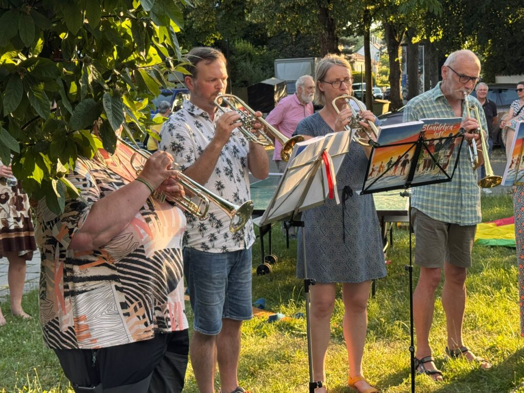 Vier Personen stehen im Freien und spielen Trompete, vor sich haben sie Notenständer mit Notenblättern. Das Sonnenlicht fällt durch die Bäume und wirft Schatten. Im Hintergrund schauen andere zu, und die Szene wirkt wie ein zwangloses Beisammensein im Freien.