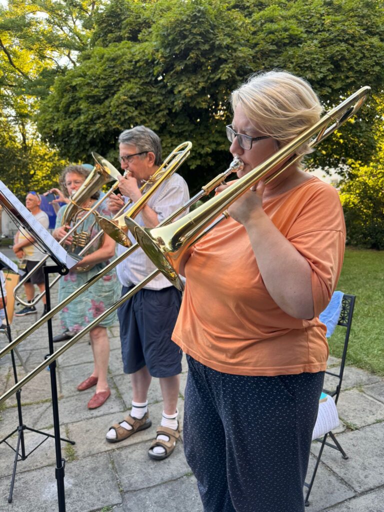 Eine Gruppe Erwachsener spielt an einem sonnigen Tag im Freien Blechblasinstrumente, darunter Posaunen. Sie stehen auf einer Steinterrasse, im Hintergrund sind üppig grüne Bäume und Gras zu sehen.