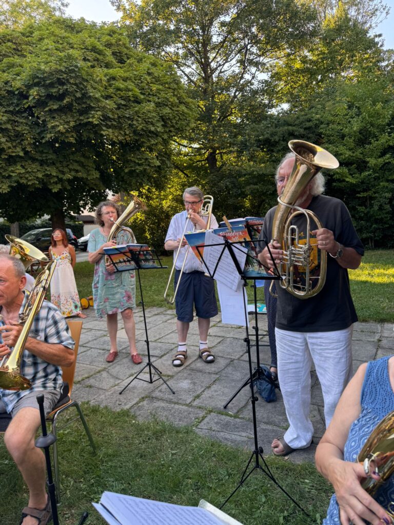 Eine Gruppe von Menschen spielt an einem sonnigen Tag im Freien Blechblasinstrumente; sie stehen oder sitzen auf Gras und Steinplatten, umgeben von Bäumen und Grünflächen, und vor sich haben sie Noten auf Notenständern.
