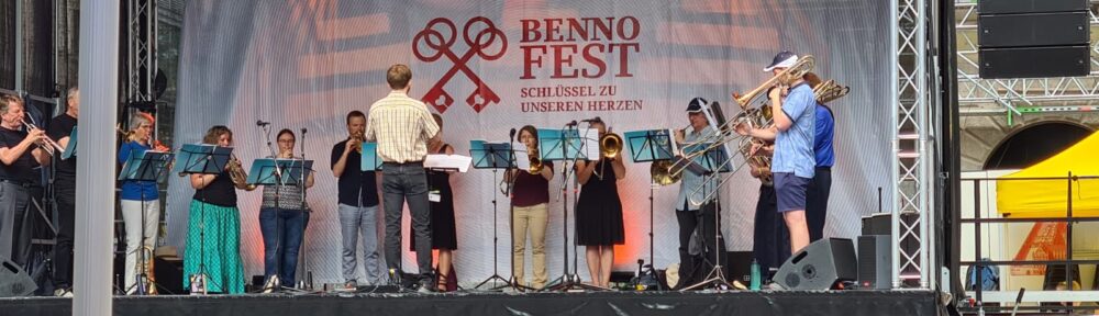 Ein Blech- und Holzbläserensemble des Posaunenchors München spielt unter der Leitung eines Dirigenten auf einer Außenbühne beim Benno-Fest. Der Bühnenhintergrund zeigt den Namen des Festivals und zwei große, gekreuzte rote Schlüssel, vor denen die Publikumsabsperrung zu sehen ist.