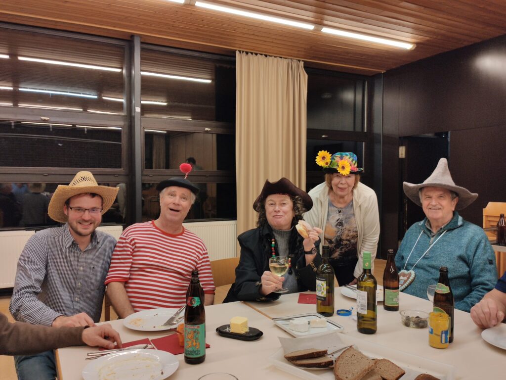 Fünf Erwachsene sitzen um einen Tisch, tragen festliche Hüte, lächeln und genießen Getränke und Essen. Die Szene sieht fröhlich aus, mit Flaschen, Tellern und Brot auf dem Tisch in einem gemütlichen, holzgetäfelten Raum.