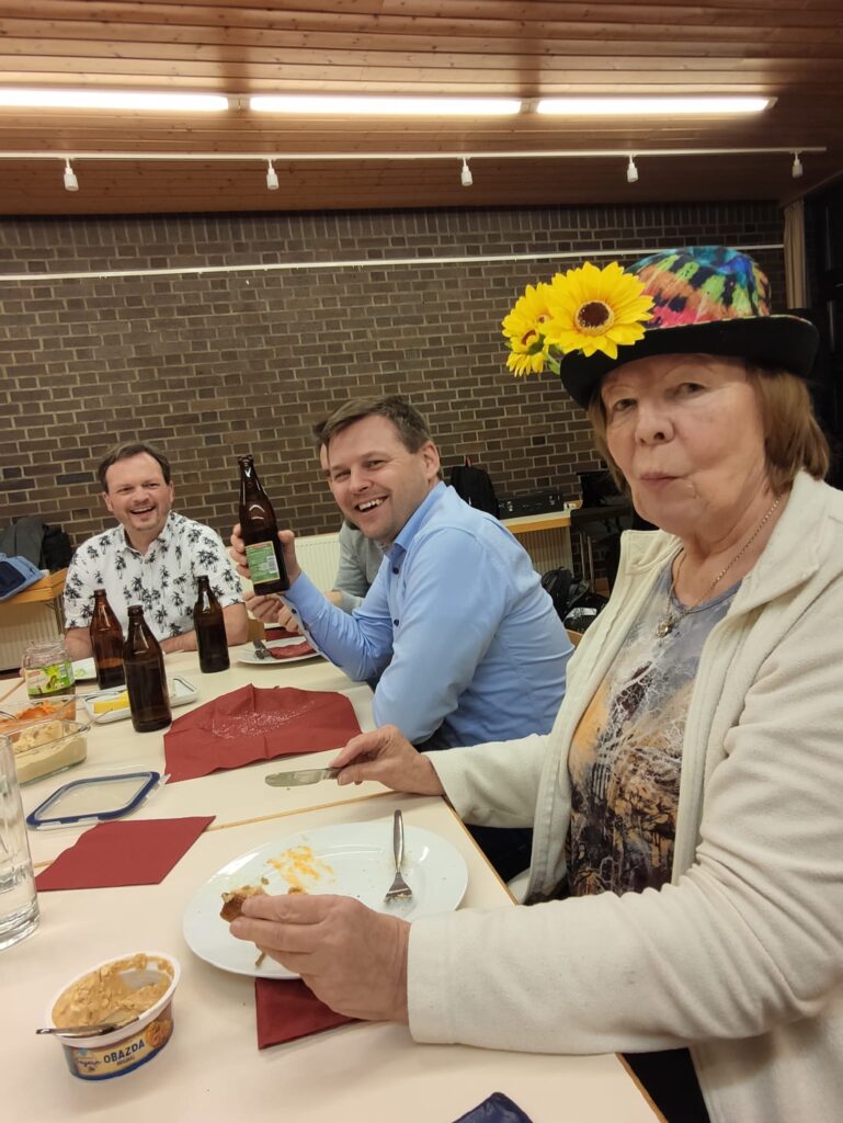 Vier Personen sitzen um einen Tisch mit Getränken und Essen. Drei Männer lächeln, einer hält eine Bierflasche hoch. Eine Frau mit einem bunten Hut mit gelben Blumen isst ein Dessert und schaut in die Kamera. Die Umgebung ist zwanglos und drinnen.