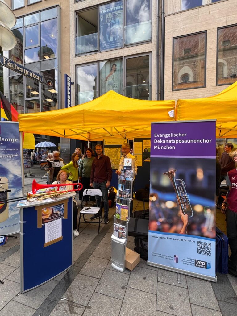 Ein Straßenfeststand mit gelben Zelten, Plakaten, einem Informationsstand und lächelnden Menschen. Auf einem Plakat wird für den Evangelischen Dekanatsposaunenchor München geworben, auf dem eine Trompete abgebildet ist. Der Stand befindet sich an einer belebten Stadtstraße.